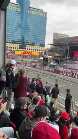 The video shows a Formula One car speeding past a crowded grandstand at a race track. The grandstand is filled with spectators, many of whom are wearing racing-themed clothing and hats. Some are holding up their phones to take pictures or videos of the race. The race track is surrounded by advertising, including signs for Qatar Airways and DHL. Tall buildings can be seen in the background.