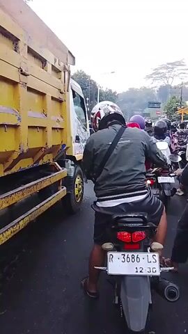 A traffic jam is depicted from the perspective of a motorcyclist. The motorcyclist is wearing a helmet and a green jacket. The camera is pointed forward, showing a yellow dump truck to the left and numerous other motorcycles in front and to the right. Some of the other motorcyclists are wearing helmets and backpacks. The scene appears to be on a road with trees and buildings visible in the background. A sign with Indonesian text is also visible.
