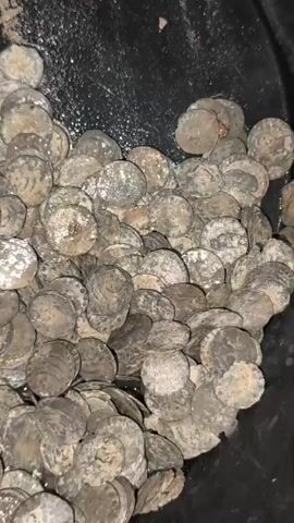 The video shows a close-up, overhead view of a pile of small, round, tarnished metal objects, likely coins, resting in a dark container. The objects are densely packed, with some overlapping. They appear to be covered in dirt or debris, giving them a dull, aged appearance. The lighting is dim, and the focus is on the texture and sheer quantity of the objects.