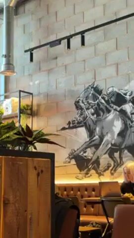The camera pans upward in a restaurant interior, focusing on a ceiling with exposed pipes, lights, and ventilation systems. Below the ceiling is a partial view of a wooden divider with plants on top, obscuring some of the view of people seated in the background. The walls are made of gray bricks and have a black-and-white mural of horses racing.