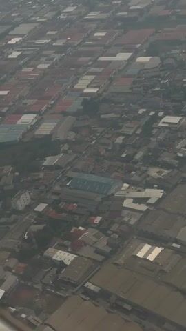 An aerial, high-angle, oblique shot pans over a sprawling industrial area filled with numerous warehouses and factories. The structures are predominantly low-rise with grey, sloped roofs, arranged in a grid-like pattern with narrow streets or pathways between them. Some roofs are lighter grey or white, suggesting different materials or solar panels. In some areas, smaller residential buildings with red or grey roofs are interspersed among the industrial complexes. The overall scene is somewhat hazy, possibly due to atmospheric conditions or pollution, obscuring distant details and giving the landscape a muted, slightly uniform appearance. There are no visible people or vehicles in the shot. The view suggests a densely populated and highly developed industrial zone.