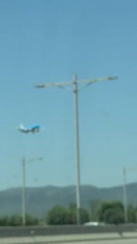 The video shows a KLM airplane flying in the sky with its landing gear down, suggesting it is either taking off or landing. The airplane is white with a blue tail and blue wingtips. It is flying in front of a clear blue sky. Below the airplane, there is a highway with cars driving on it. There are also some trees and hills in the background. The video is shot from a moving vehicle, likely a car, as indicated by the blurred foreground and the perspective. The scene appears to be at an airport or near an airport, given the presence of the airplane and the highway.
