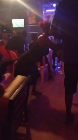 The video shows a woman dancing in a dimly lit club or bar. She is wearing a black dress and has her hair up. She is bent over and appears to be dancing suggestively. There are other people in the background, some of whom are also dancing or sitting. The environment is decorated with lights and there is a television screen visible. The music is not audible in the video.