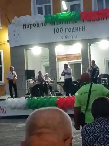 This video shows a band performing on a stage in front of a building. The building has a banner that reads "NARODNO CHITALISHTE 100 GODINI s. Konush," which translates to "Community Center 100 Years s. Konush." The banner is decorated with balloons in the colors of the Bulgarian flag: white, green, and red. The band consists of several musicians playing various instruments, including a saxophone, an accordion, and what appears to be a clarinet or a similar wind instrument. A keyboard or synthesizer is also visible on the stage. There are speakers and other audio equipment. Several people are watching the performance, and some are standing in the foreground, facing away from the camera. The lighting suggests it is either dusk or evening.