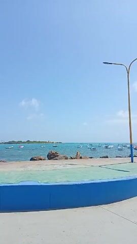 The video shows a sunny day at the beach from what appears to be a slow-moving vehicle. The sea is visible in the background, and several small boats are on the water. A small island is visible on the horizon. In the foreground, a blue and green platform sits between the camera and the water's edge. There are some rocks lining the shore. As the vehicle continues forward, yellow streetlights come into view, and a palm tree is seen. The sky is blue with a few clouds.