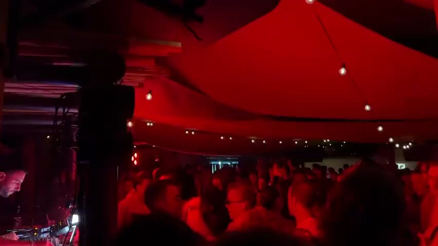 A crowded nightclub with red lighting. A DJ is visible on the left, mixing music. People are dancing and enjoying themselves. The ceiling is draped with red fabric, and string lights are hung across it.