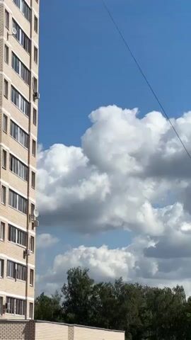 The video begins with a vertical shot of a tall, beige apartment building on the left side. The building has multiple floors with visible windows and air conditioning units. The sky above is a bright blue with fluffy white clouds. The middle of the video shows the apartment building from a slightly different angle, with a row of cars parked in front of a smaller building or entrance. Trees are visible in the background. The video then shifts to a shot of utility poles with wires stretching across the frame against the blue sky and clouds. The final shot shows the leaves of a tree against the sky, with clouds partially visible. The video appears to be filmed outdoors on a sunny day.