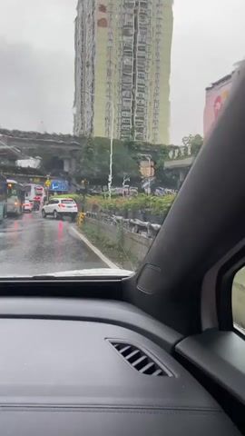 The video shows the view from inside a car driving on a wet road in what appears to be a city. The car's dashboard is visible in the foreground, and the windshield has raindrops on it, indicating it is raining. The side-view mirror on the right side of the car is also visible, showing raindrops and a reflection of the road and surroundings. In the background, there are buildings, including a tall, light-colored apartment building and a lower, white building with a large mural of a heart. There are also cars on the road, including a white car ahead. The road appears to be an elevated highway or overpass, with infrastructure supporting it visible. There are also plants and flowers in planters along the side of the road. The overall impression is a rainy day drive through an urban environment.