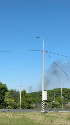 The video shows a rural scene with a street and a field, with a lamppost in the foreground. Behind the trees and in the distance, there is a gray cloud of smoke.