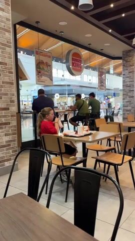 The video shows the interior of a Burger King restaurant with several people present. In the foreground, a young woman with blonde hair in a ponytail, wearing a red shirt and black shorts, sits at a table with a tray containing a drink and food wrappers. She is holding a cup and appears to be drinking. In the background, a man in a dark shirt stands near a glass door, looking outward. Two men in green shirts sit at a high table, facing away from the camera, with food in front of them. The restaurant has a modern interior with brick and wood accents. Through the large glass windows, a shopping mall or public space with other shops and people can be seen. The Burger King logo is visible hanging from the ceiling. The overall lighting suggests daytime.