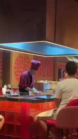 The video shows a teppanyaki-style restaurant setting, with a chef in a purple uniform preparing food on a flat grill in front of a counter where people are seated. The patrons are facing the chef and watching him cook.  The restaurant has a red color scheme with red stools, red tile, and decorative red lights on a shelf.  The ceiling is a dark grid pattern with decorative orange and yellow lamps hanging down. The restaurant appears busy with multiple customers seated.