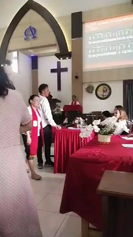 In a church setting, a group of people are gathered. A woman in a pink polka-dot dress is walking away from the camera towards the left side of the frame. A man in a white shirt and a woman in a white shirt with a red sash are walking towards the right side of the frame. Another woman in a patterned dress is standing near a wooden structure on the left. The church has a cross on the wall and a projector screen displaying text. Red tablecloths are visible on tables.