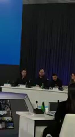 The video captures an indoor scene at what appears to be a conference or meeting in Barvinok, Ukraine. Several individuals are seated at a long white table, facing forward. At least four people are visible in the midground, including Ukrainian President Volodymyr Zelenskyy (center right, in black). Microphones and water bottles are placed on the table in front of them, along with what appear to be laptops. The background features a dark blue curtain, possibly a stage backdrop, and a bright blue screen or wall to the left. In the foreground, the back of a person's head and shoulder are visible, suggesting an audience perspective. The people at the table appear to be listening attentively, possibly to a speaker who is not fully visible in the frame, or engaged in a discussion. No specific emotions are clearly discernible, but the overall atmosphere seems focused and professional. There is no visible text in the scene.