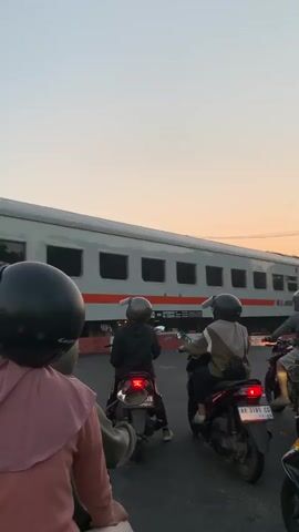 A wide, ground-level outdoor shot shows a group of people on scooters stopped at a train crossing. The train is moving from left to right, and the sun is setting in the background. The camera is still, and no one is moving. The train is grey with an orange stripe, and "KAI" is written on the side.