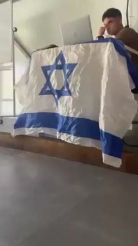 The video shows a person sitting at a desk covered with an Israeli flag and watching a news broadcast on TV. The news anchors are reporting on a war, with the text “טראמפ: זה יום גדול, המלחמה נגמרה” visible on the screen. The video appears to be taken from a low angle.
