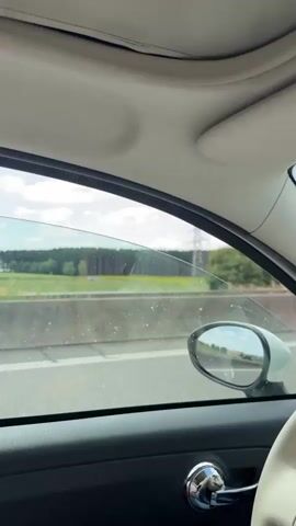 The video is filmed from inside a car, looking out the passenger window as it travels along a highway. The car interior is visible in the foreground, including the door panel, door handle, and part of the ceiling. The window is partially lowered, allowing a view of the outside environment. Outside, a highway passes by, with a concrete barrier separating the lanes. Beyond the highway, there are fields with yellow flowers and green grass, bordered by a dark green forest in the distance. The sky is blue with some scattered clouds. In the middle distance, a plume of dust or smoke is visible, being blown across the field. A power line pylon is also visible in the background. The side mirror reflects the passing scenery. The video appears to be a continuous shot, capturing the movement of the car and the changing landscape.