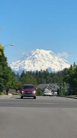 The video begins with a wide shot of a snow-capped mountain dominating the background under a clear blue sky. In the middle ground, a dense line of green trees forms a natural border below the mountain. In front of the trees, several suburban houses with dark roofs are visible. A red SUV is driving towards the camera on an asphalt road that occupies the foreground. The SUV is in motion, and its headlights are on. To the left of the road, there is a sidewalk and a short fence. A streetlight is visible on the left side of the frame. To the right of the road, there is another house and a speed limit sign that reads "25". Another vehicle, possibly a lighter-colored SUV, is parked in the driveway of one of the houses. The scene appears to be a residential neighborhood on a sunny day.