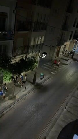The video shows a street scene at night. There are buildings lining the street, some with lights on. Several people are visible, walking and standing. There are also trees and streetlights. The street is paved and appears to be cobblestone. In the foreground, there is a balcony or railing. The camera appears to be mounted on a drone or elevated platform, as the view is from above. There is some motion blur throughout the video, indicating movement of the camera or objects within the scene. Some vehicles are visible in the background, including a car and a scooter. There is a pedestrian crossing marked on the street. A trash can is visible near the sidewalk.
