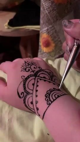 A close-up shot shows a hand with a black henna design being applied to the back of the hand and wrist. The henna is being applied from a cone-shaped applicator by a person's right hand, which is holding the cone and guiding it. The design features intricate patterns including swirls, dots, and a floral motif. The hand receiving the henna is resting on a light-colored fabric. In the blurred background, there appears to be a table with a dark object and a decorative item with a sunflower pattern. The lighting is dim, suggesting an indoor setting.