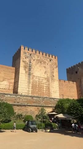 The video captures a sunny day at the Alhambra in Granada, Spain, as evidenced by the bright blue sky and strong shadows. The main subjects are the impressive ancient fortress walls and towers, constructed from warm-toned stone and featuring crenellated battlements. In the foreground, a well-maintained garden area with lush green bushes and trees provides a stark contrast to the arid, historical architecture.

Several people are visible in the scene. On the left, a person walks on a dirt path, seemingly exploring the grounds. Near the center, a small, dark-colored utility vehicle, possibly used for maintenance or transport within the complex, is parked. To the right of the vehicle, a group of people are gathered under two large beige umbrellas, suggesting a rest area or a meeting point. Further to the right, another group of people stands together, some looking towards the fortress, others interacting amongst themselves. The overall atmosphere appears relaxed and leisurely, typical of a tourist destination.

Several dark birds are seen flying in the clear sky, adding a touch of life to the scene. The architecture of the Alhambra, with its weathered stone and imposing scale, dominates the background, evoking a sense of history and grandeur. The time of day is clearly midday, given the high sun and the shadows cast. There is no visible text in the video.
