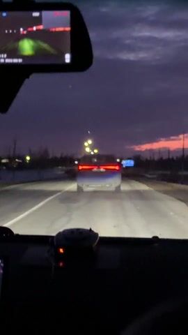 A vehicle is traveling on a road, following another car. It's twilight, with a colorful pink and purple sky visible. There's snow on the ground alongside the road, and power lines can be seen silhouetted against the sky. The view is from inside the car, looking through the windshield, with a rear-view mirror and dashboard visible.