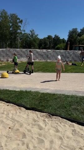 In a sunlit outdoor area, a young child wearing a light-colored swimsuit and a white head covering runs across a sandy patch in the foreground. Behind the sand, a paved walkway stretches across the middle ground. On the walkway, two children are riding scooters. The child on the left is wearing a green shirt and helmet, while the child on the right is wearing a dark shirt. Further back on the walkway, a man stands next to a small motorbike with two children on it, one appearing to be riding and the other a passenger. In the background, a large yellow construction vehicle is visible behind a temporary fence with red and white markings. Trees and a clear blue sky complete the scene.