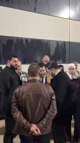 A group of about a dozen people stand closely together, talking, indoors. Most are men dressed in business-casual attire, including suits, jackets, and dress shirts. At least one woman is present, wearing a white head covering and a dark coat. The people are clustered near a wall which is decorated with a large, dark, abstract design. The camera moves slightly, and the faces of some individuals are partially visible. One man with a beard seems to be the focal point of the conversation. A large man in a black suit stands at the edge of the group. The overall impression is a business meeting or an arrival scene where people are conferring.