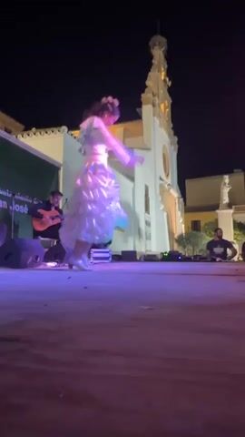 A group of people are on a stage at night in front of a building with a steeple. A woman in a ruffled dress is dancing. A man is playing a guitar. Another man is sitting behind a table with equipment. The stage is lit with purple lights.