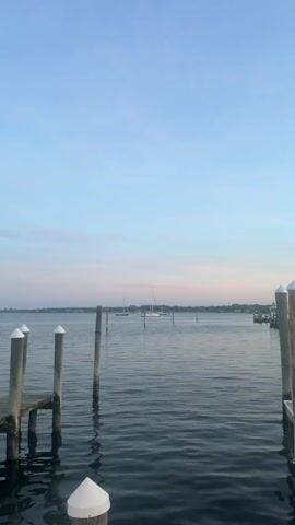 The video shows a calm body of water with a clear blue sky transitioning to soft pinks and oranges near the horizon. Several wooden posts, some with white caps, rise from the water, indicating a dock or pier. In the distance, sailboats are anchored on the water. The water is still, with gentle ripples reflecting the soft light of the sky. The overall impression is one of peace and tranquility, possibly at sunrise or sunset.