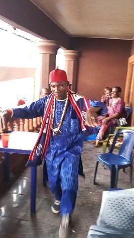 The video shows a man in a blue robe and red hat dancing on a patio with pillars. He moves toward a group of women sitting in chairs. The man kneels on the floor and hugs one of the women. The setting is outdoors, but the patio is covered. There is a table with items on it in the background. Some red plastic cups are visible.