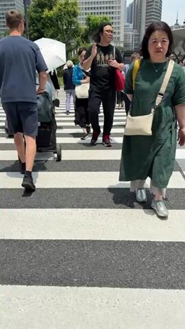 The video shows a pedestrian crossing with several people walking across the street. In the foreground, a person in a green dress is partially visible on the right. Further back, a man wearing a black t-shirt and black pants, with a red bag over his shoulder, walks with his hand raised. Behind him, other pedestrians are crossing the street, some carrying umbrellas. A woman in a light-colored dress and white cardigan is also visible, holding a black umbrella. Further back, a man in a navy blue t-shirt and shorts is pulling a suitcase. In the background, there are buildings and traffic lights. The overall scene depicts a busy urban environment with pedestrians navigating a crosswalk on a sunny day.