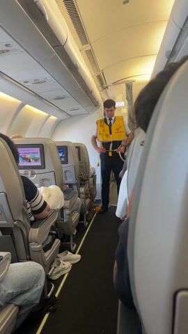 The video shows a flight attendant demonstrating how to put on a life vest. He is standing in the aisle of an airplane, facing the camera. He is wearing a white shirt, black tie, black pants, and a yellow life vest labeled "DEMO ONLY." He is adjusting the straps of the life vest. Passengers are seated on either side of the aisle. The interior of the plane is visible, including the seats, overhead bins, and video screens. The lighting is bright and artificial.