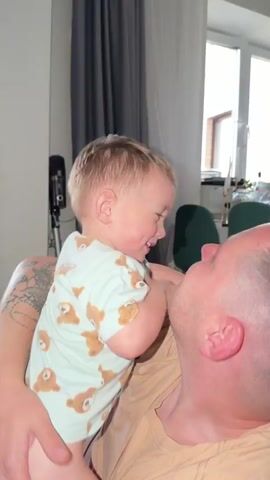 A toddler with blonde hair and a light green t-shirt with a bear pattern is being held by a man. The toddler is reaching out and touching the man's face, specifically his nose and mouth, and appears to be laughing. The man is looking up at the toddler and has a relaxed expression. They are indoors, with a window in the background showing a brick building and trees. A dark curtain is visible to the left.