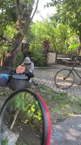 The video shows a rusty, vintage bicycle with a large woven basket attached to the rear, filled with items. The bicycle is parked on a cobblestone pathway surrounded by lush green foliage. The scene appears to be outdoors, possibly in a residential area or a village, given the presence of trees and a somewhat weathered structure in the background. The camera is positioned at an angle, looking towards the bicycle.