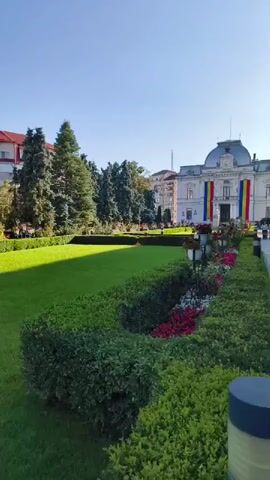 The video shows a sunny day with a clear blue sky over a city. In the foreground, there is a well-maintained garden with lush green grass and meticulously trimmed hedges. Colorful flowers, predominantly red and white, are planted along a border. Further back, several tall evergreen trees stand on the left side of the frame. Behind the trees and the garden, a large, ornate white building with a domed roof is visible. The building is decorated with large vertical Romanian flags. A paved walkway is on the right side of the frame, and a modern lamp post is in the lower right corner. People can be seen walking in the distance near the building, but they are not the focus of the video. The overall impression is one of a clean, organized, and festive public space.
