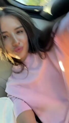 A short video shows two young women in the back seat of a car. The woman on the right, with long dark hair, wears a pink sweatshirt and makes a kissing face at the camera. The woman on the left, wearing glasses and a white sweatshirt, is partially visible and blurred, looking upwards. The video is shaky and appears to be filmed by one of the passengers. The interior of the car, including seats and windows, is visible. Sunlight streams in through the windows, creating bright highlights and shadows. The overall mood is casual and playful.