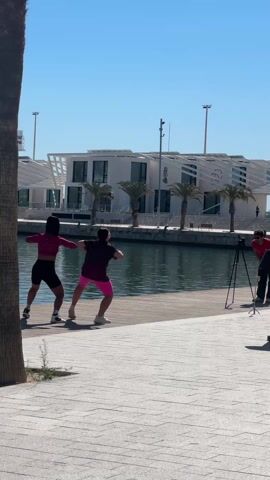 Two people are dancing on a wooden walkway near a body of water, while a person films them with a camera on a tripod. The dancer on the left is wearing a pink long-sleeved shirt and black shorts, while the dancer on the right is wearing a purple t-shirt and pink shorts. They are performing in unison, facing away from the camera. Behind them, across the water, is a modern white building with palm trees in front. The sky is blue and clear. A palm tree trunk is visible on the left side of the frame. The walkway is paved with grey patterned tiles.