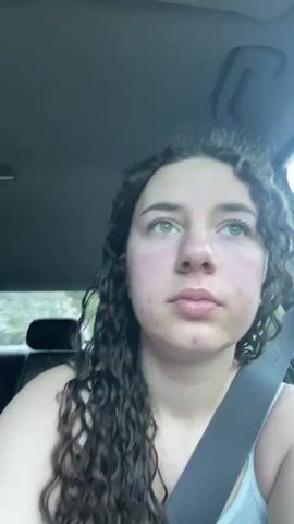 The video shows a young woman with curly brown hair sitting in the driver's seat of a car. She is wearing a gray tank top and a seatbelt. The car's interior is visible. Initially, she has a neutral expression. She then smiles at the camera, before exaggerating her lips in a "fish face" expression. The background is blurred, suggesting the car may be in motion or the focus is primarily on the woman.