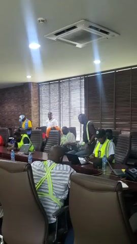 The video shows a group of people in what appears to be a conference room. Most of the individuals are wearing high-visibility vests in yellow and green, suggesting they are involved in some sort of work or training. Some are seated at a large conference table, while others are standing. Laptops and water bottles are visible on the table. The room has a brick-patterned wall and large windows covered by blinds. There are overhead lights and an air conditioning unit visible on the ceiling. Some individuals appear to be speaking or listening attentively. The overall atmosphere seems to be a meeting or presentation.