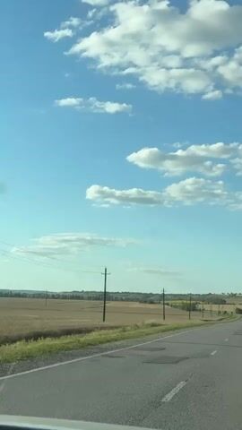 A car travels down a rural road on a sunny day with scattered clouds. The road is asphalt and shows signs of wear with several patched potholes. On either side of the road are fields of crops, possibly wheat and corn, with utility poles and power lines running along the roadside. In the distance, rolling hills can be seen under a bright blue sky. The car is white and is traveling away from the camera.