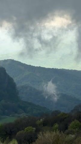 The video shows a landscape view of mountains covered in dense green forest under an overcast sky. The sky is filled with dark, heavy clouds, creating a dim, hazy atmosphere. Low-hanging clouds or mist are visible in the valleys and around the mountains, obscuring parts of the forest. The foreground features trees and shrubs, providing a sense of depth to the scene. The overall mood is subdued and atmospheric.
