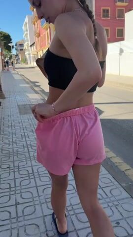 A young woman walks down a patterned sidewalk in a sunny, outdoor setting. She wears a black strapless top and pink shorts, with a glimpse of brown underwear. Her hair is in a braid. She carries a bag and adjusts her shorts. The street is lined with buildings.