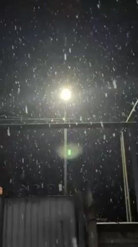The video shows a night scene with heavy snowfall. A street lamp illuminates the falling snow, creating a stark contrast against the dark background. The snow appears to be large, wet flakes. A metal fence is visible in the foreground.  Someone can be heard speaking in the background.