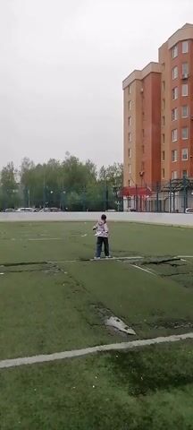 A person is walking and flailing two white objects, seemingly sticks or poles, on a green turf field. The field has white lines and some areas of damage or discoloration. Behind the field is a tall brick building with multiple windows and some trees. The sky is overcast. The person is wearing a patterned jacket and dark pants. They are moving in a somewhat erratic manner.