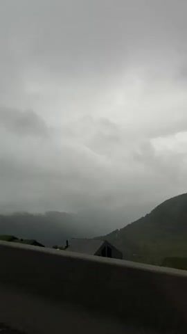 This media file captures a view from a roadside, likely a mountain pass, in Røldal, Norway. The scene is dominated by a dramatic, overcast sky filled with heavy, gray clouds that obscure the sun, suggesting a gloomy or stormy day. Rolling, mist-shrouded mountains stretch into the distance, their green slopes softened by the atmospheric conditions.

In the foreground, a dark, concrete barrier runs horizontally across the lower portion of the frame, suggesting the perspective is from a vehicle or a lookout point. A few scattered buildings with dark roofs are visible nestled on the slopes of the mountains, appearing small against the vast landscape. One building in particular, with a peaked roof, is more prominent in the middle ground. The overall atmosphere is one of isolation and raw, natural beauty, amplified by the moody weather. There is no visible text within the media file. The time of day is unclear due to the cloud cover, but the mood suggests it could be late afternoon or evening, or simply a day where the weather is consistently overcast. The scene evokes a sense of quiet contemplation and the power of nature.