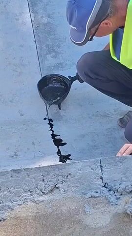 A person wearing a blue cap, glasses, and a yellow safety vest is kneeling and pouring black liquid from a dark-colored container onto a gray concrete surface with a crack. The liquid is thick and viscous, forming a wavy line as it flows into the crack. The person is holding the container with a gloved hand and tilting it to pour. The background shows more of the gray concrete surface. The scene appears to be outdoors on a sunny day.