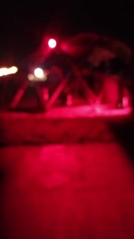The video depicts a night scene with a red light illuminating a sandy area where a metal structure is located. This structure appears to be a tent-like construction with multiple supporting beams, inside which several people are visible. They are standing and engaging in activities that are difficult to discern clearly due to the darkness and the red lighting. The overall setting suggests an outdoor gathering or event. The foreground consists of a paved surface, also bathed in red light, providing a path leading towards the structure. The video is somewhat shaky and the details are not sharp.