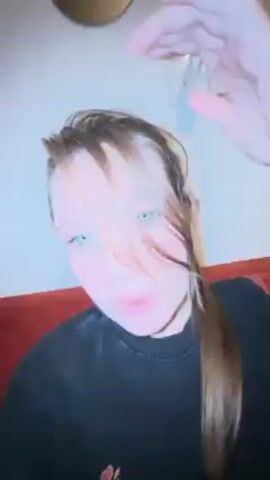 A close-up shot shows a young woman indoors. She has light brown hair pulled into a ponytail. She lifts her hair to show her face, which has fair skin and green eyes. The woman wears a black t-shirt with a red design on the left chest. The background includes a white wall with an electrical outlet and a red couch or cushion.