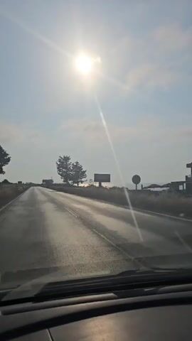 A video captures a view from inside a car driving down a highway on a sunny day. The sun is bright and high in the sky, casting lens flares across the image. The road ahead is empty, with lanes marked by white lines. On either side of the road, there are trees and some distant structures, possibly buildings or signs. The sky is blue with some scattered clouds. The overall impression is of a scenic drive through a coastal or rural area.