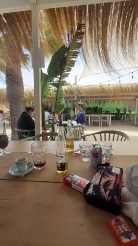 The video shows a table at an outdoor restaurant. There are several drinks and a pack of cigarettes on the table. In the background, people are seated at other tables, and palm trees and thatched roofs are visible. The scene appears to be a beachside establishment on a sunny day.